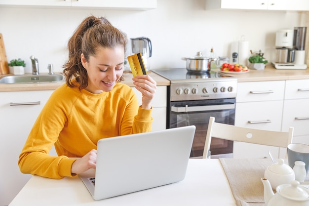 Donna che compera online e paga con carta di credito oro. Giovane ragazza seduta con laptop acquisto su Internet immettere i dettagli della carta di credito su sfondo cucina interna.