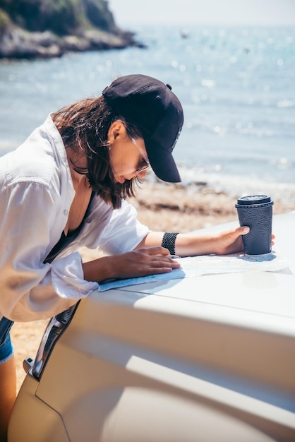 Donna che controlla la mappa sul cofano dell'auto che beve caffè al concetto di viaggio sulla spiaggia del mare d'estate