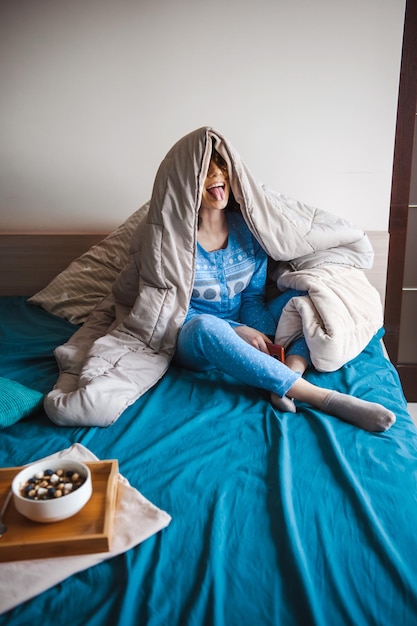 Donna che copre la testa con una coperta seduta a letto mentre fa una smorfia mattutina mangiando sano