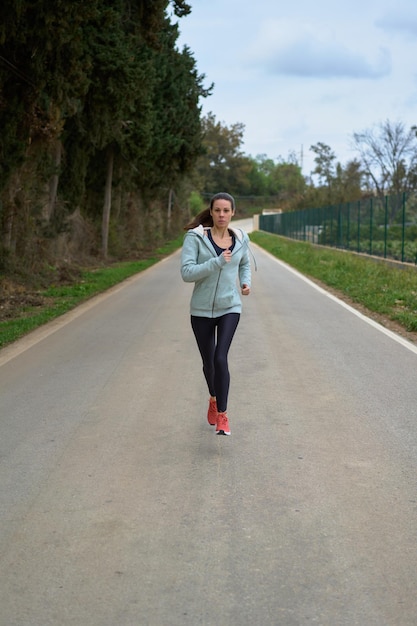 Donna che corre su una strada asfaltata attraverso la vista frontale della foresta con leggings e felpa