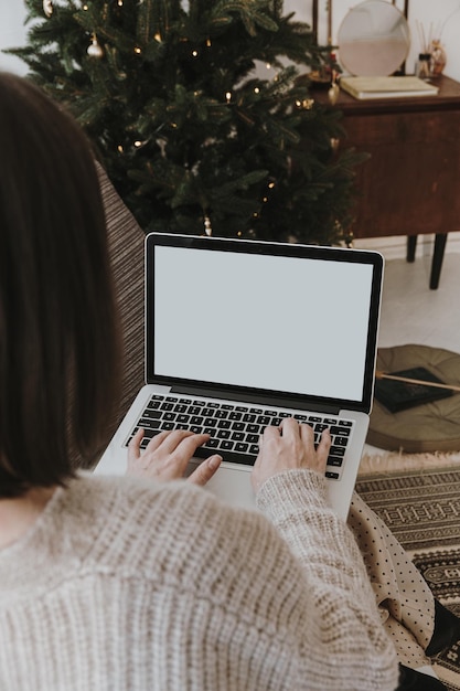 Donna che digita su un computer portatile con schermo vuoto in un confortevole e accogliente soggiorno interno decorato con albero di Natale