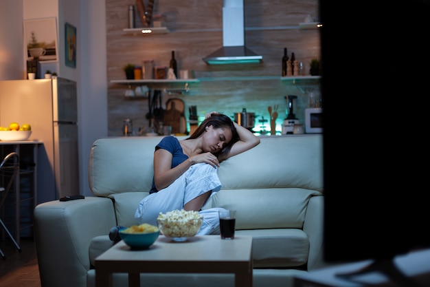 Donna che dorme su un divano davanti alla TV mentre guarda un film annoiato. Stanca esausta solitaria donna assonnata in pigiama che si addormenta seduta su un comodo divano in soggiorno, chiudendo gli occhi di notte