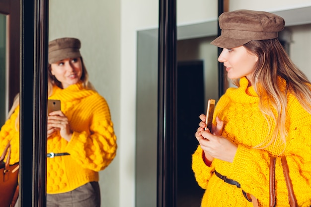 Donna che esamina specchio a casa indossando maglione ed accessori alla moda e prendendo selfie