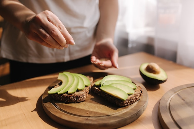 Donna che fa panino vegano utilizzando avocado pane di segale e semi di sesamo. Donna cospargere di sesamo