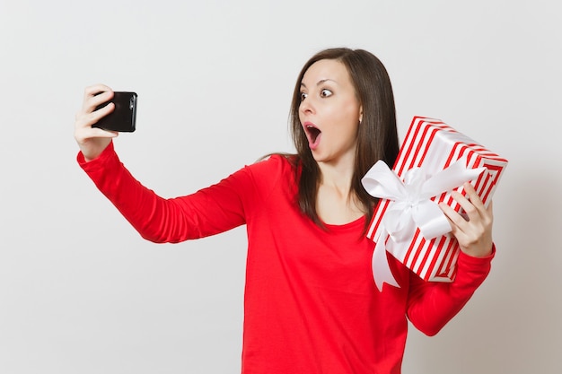 Donna che fa selfie sul telefono cellulare con scatola regalo a strisce rosse con regalo isolato su sfondo bianco. Copia spazio per la pubblicità. San Valentino, Giornata internazionale della donna, concetto di vacanza.