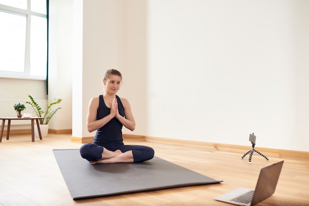 Donna che fa yoga online. Computer e fotocamera nel suo salotto. Stile di vita sano e concetti di lavoro a distanza