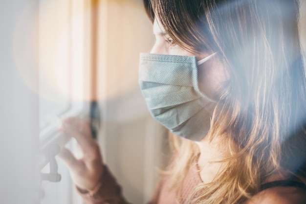 Donna che guarda fuori dalla finestra con una maschera chirurgica durante il blocco per il coronavirus covid-19