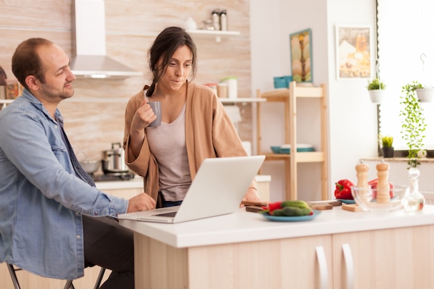 Donna che guarda il laptop del marito mentre sta lavorando in cucina. Fidanzata che tiene tazza di caffè. Felice amorevole allegro romantico innamorato coppia a casa utilizzando la moderna tecnologia internet wireless wifi