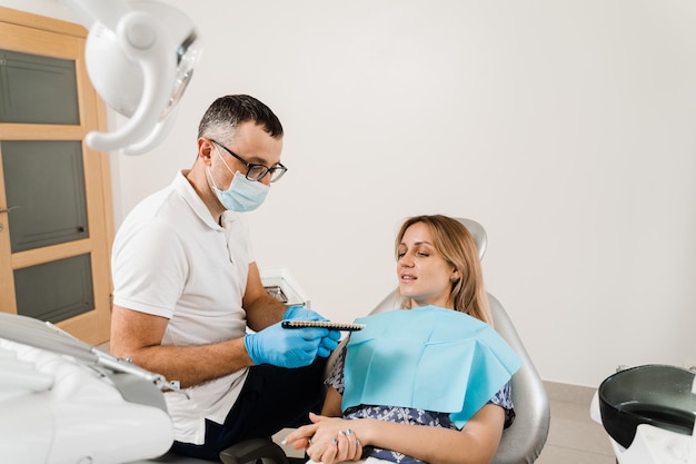 Donna che guarda le faccette o gli impianti campioni di corrispondenza del colore dei denti nelle mani del medico Odontoiatria Il dentista dimostra la guida alle sfumature di colore dei denti per lo sbiancamento dei denti per la donna paziente in clinica odontoiatrica