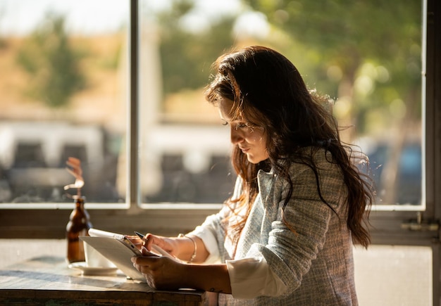 Donna che guarda lo schermo del tablet digitale mentre è seduto in un bar accanto alla finestra