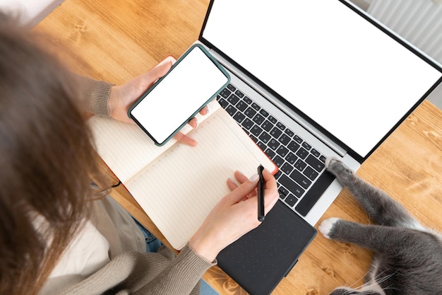 Donna che guarda sullo schermo dello smartphone e prende appunti in un blocco note su un computer portatile da tavolo in legno