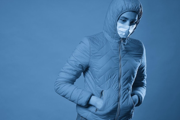 Donna che indossa il cappuccio in una maschera protettiva e cappotto su sfondo blu Modello ragazza in maschera medica e abito primaverile girato in studio con copia spazio per il tuo testo Antiepidemico antiinfluenzale