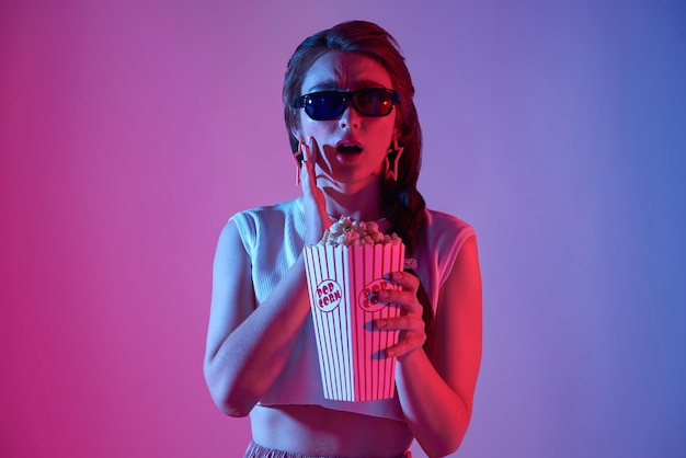 donna che indossa occhiali 3D con popcorn e bevande guardando film emozionanti su sfondo neon