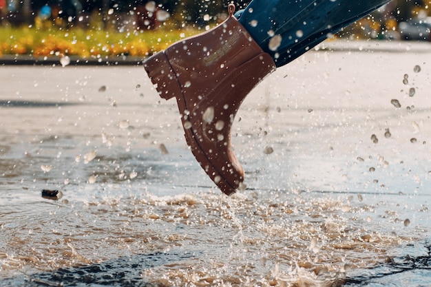 Donna che indossa stivali di gomma da pioggia camminando correndo e saltando in una pozzanghera con spruzzi d'acqua e gocce sotto la pioggia autunnale