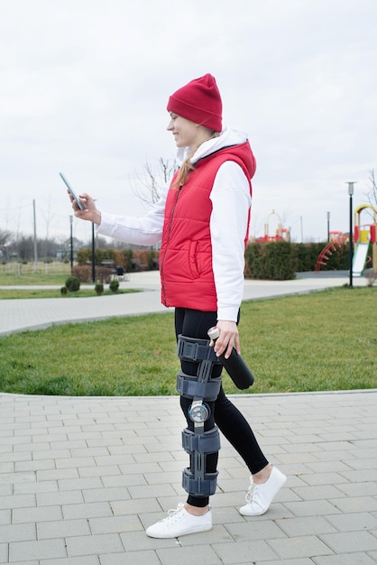 Donna che indossa tutore per ginocchio o ortesi dopo un intervento chirurgico alle gambe che cammina nel parco utilizzando lo smartphone