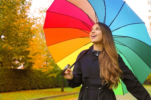 Donna che indossa un impermeabile scuro sotto l'ombrello arcobaleno nel parco cittadino