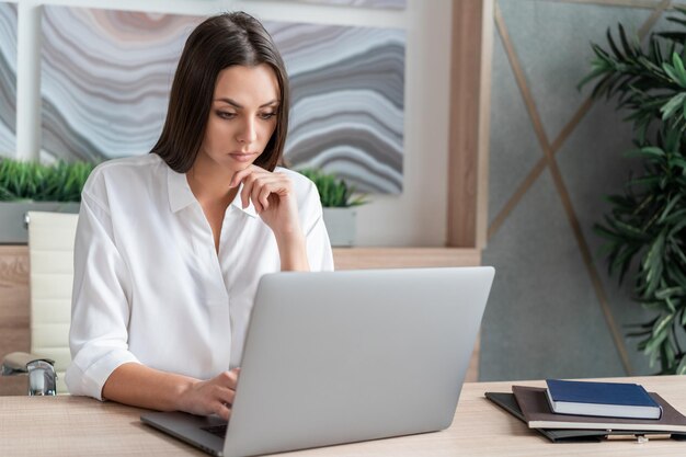 Donna che indossa una camicia bianca seduta Tavolo con laptop e notebook per prendere appunti Concetto di lavoro