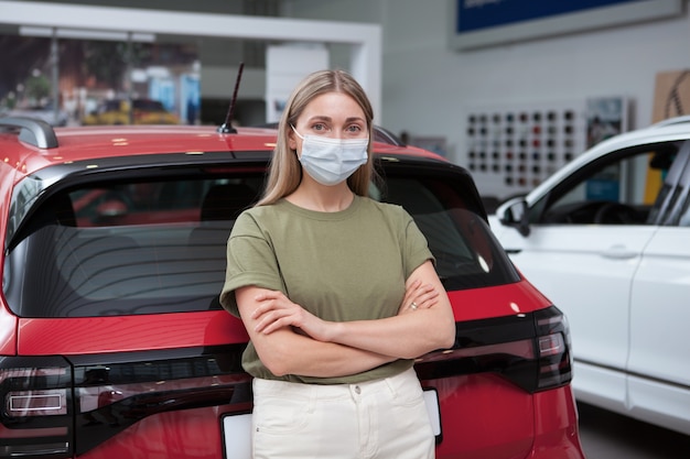 Donna che indossa una maschera medica, appoggiata a una nuova auto in una concessionaria di auto