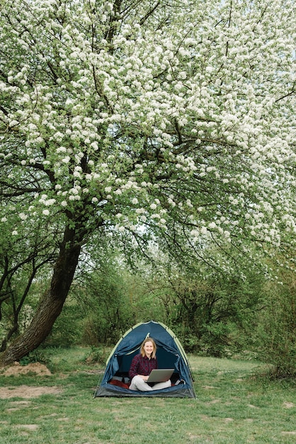 Donna che lavora al computer portatile in tenda in natura. Giovane professionista che si siede nell'accampamento. Rilassarsi in campeggio nella foresta, prato. Lavoro a distanza, attività all'aperto in estate. Ragazza felice che si distende, lavora in vacanza.