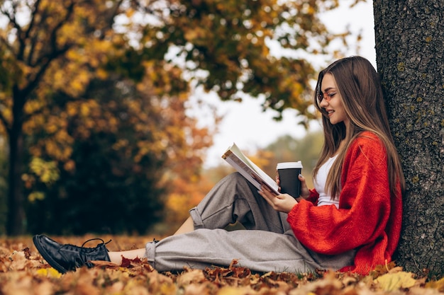 Donna che legge in un parco autunnale e beve caffè sotto l'albero