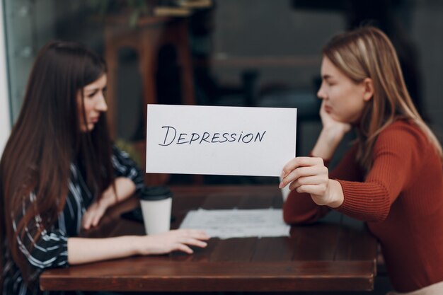 Donna che mantiene il foglio bianco di carta con etichetta parola depressione in mano. Due donne che parlano in un caffè di strada. Concetto di depressione nascosta.