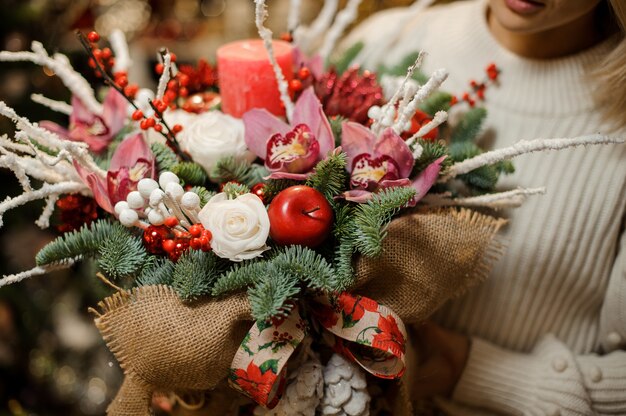 Donna che mantiene una composizione natalizia con orchidee rosa, rose bianche, rami di abete, mela rossa e candela in tela di sacco nel negozio di fiori