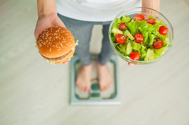 Donna che misura il peso corporeo sulla bilancia tenendo hamburger e insalata