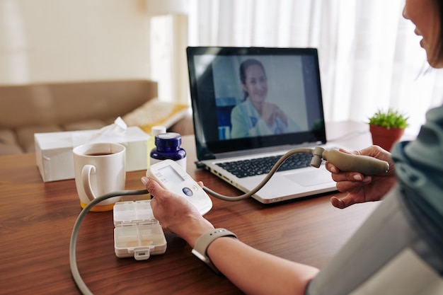Donna che misura la sua pressione sanguigna con il tonometro elettronico a casa sotto il controllo del medico virtuale