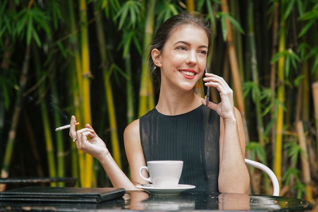 Donna che parla al suo telefono, fumando e mangiando caffè