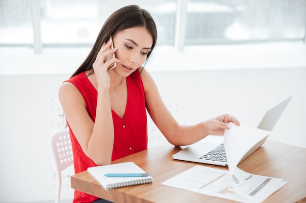 Donna che parla al telefono e si siede al tavolo con il laptop in ufficio