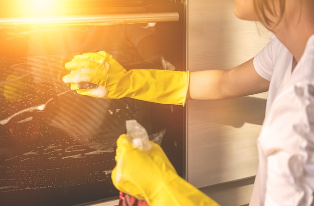 Donna che pulisce il forno in cucina, mano in guanti di gomma gialli con straccio e detersivo spray, foto di sfondo del concetto di lavoratore di servizio