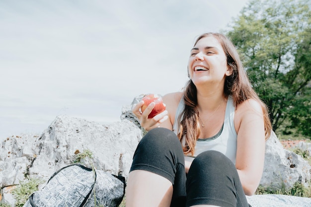 Donna che ride mentre mangia una mela in montagna, lungo sentiero, attività escursionistica, tempo libero e sport naturale