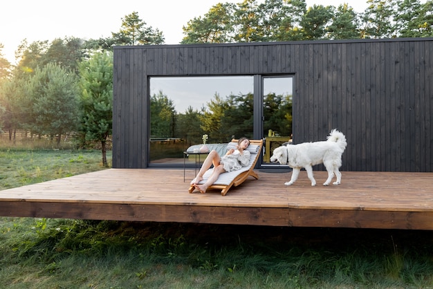 Donna che riposa sul lettino sulla terrazza in legno vicino alla casa moderna con finestre panoramiche vicino alla pineta con un grosso cane bianco. Benessere e salute consapevole. Copia spazio