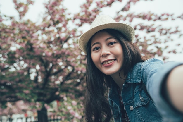 Donna che scatta foto di sé nella stagione primaverile con un albero di ciliegio rosa. giovane ragazza viaggiatore indossare cappello sorridente fotocamera faccia facendo selfie con il fiore di sakura nel parco all'aperto di osaka giappone. ritratto femminile asiatico