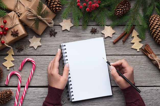 Donna che scrive la lettera di Natale o gli obiettivi del nuovo anno su carta su sfondo di legno con decorazioni, vista dall'alto, spazio di copia