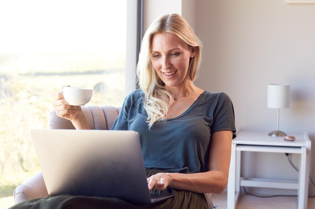 Donna che si distende sulla sedia dalla finestra a casa usando il computer portatile che beve caffè