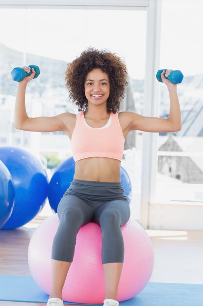 Donna che si esercita con i dumbbells sulla sfera di forma fisica in ginnastica