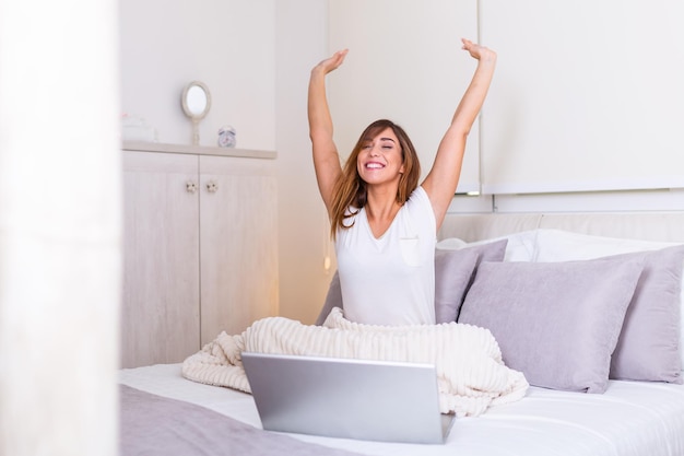 Donna che si estende a letto dopo essersi svegliata al mattino pronta per lavorare da casa. Ritratto di giovane donna sorridente che si siede con il computer portatile sul letto. Concetto di lavoro freelance da casa