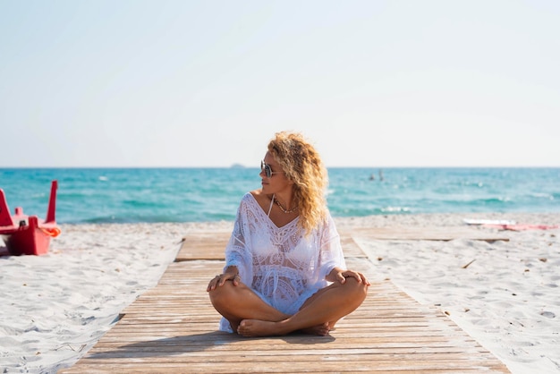 Donna che si gode il sole e l'abbronzatura in spiaggia in estate vacanza viaggio vacanza attività per il tempo libero Una donna seduta su un molo di legno sulla sabbia con oceano blu sullo sfondo Turistico di destinazione panoramica