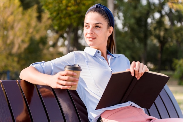 Donna che si siede su una panca di legno, leggendo un libro e bevendo caffè