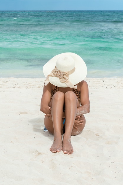 Donna che si siede sulla sabbia che abbraccia le sue gambe. Donna in costume da bagno e cappello sulla spiaggia. Swell. Donna abbronzata Abbronzatura. Concetto di vacanza e relax