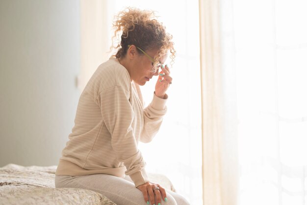 Donna che soffre di stress o mal di testa che fa smorfie di dolore seduta sul letto in camera da letto a casa al mattino