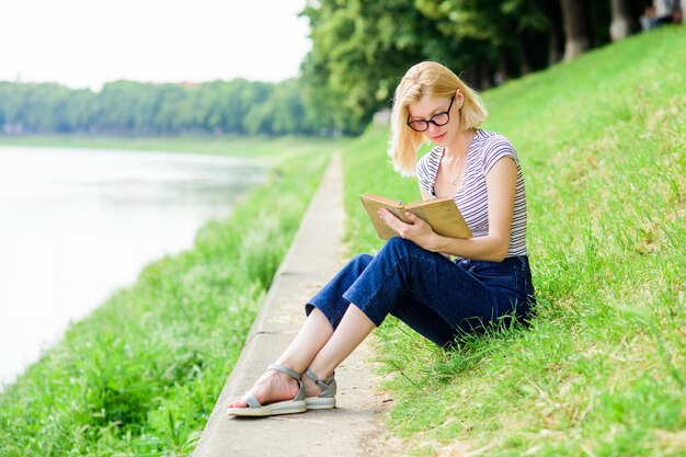 Donna che sogna le vacanze Letteratura per le vacanze estive Riposo relax e hobby Vacanze estive La ragazza si siede sull'erba verde vicino al fiume e legge il libro Ragazza che si rilassa in riva al fiume dopo una giornata lavorativa