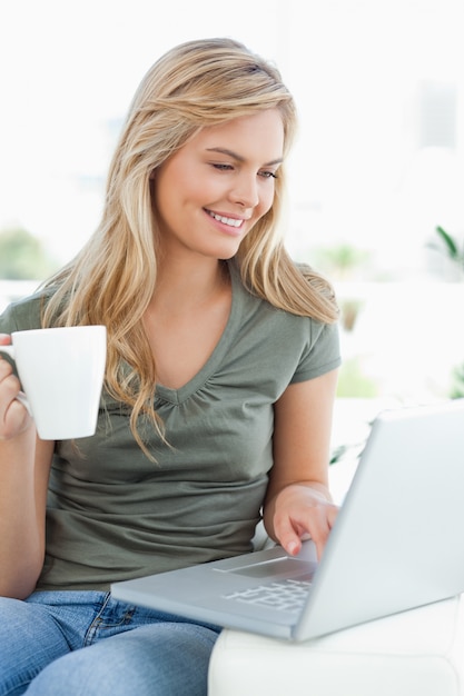 Donna che sorride, mentre usa il suo laptop e tiene una tazza nell&#39;altra mano