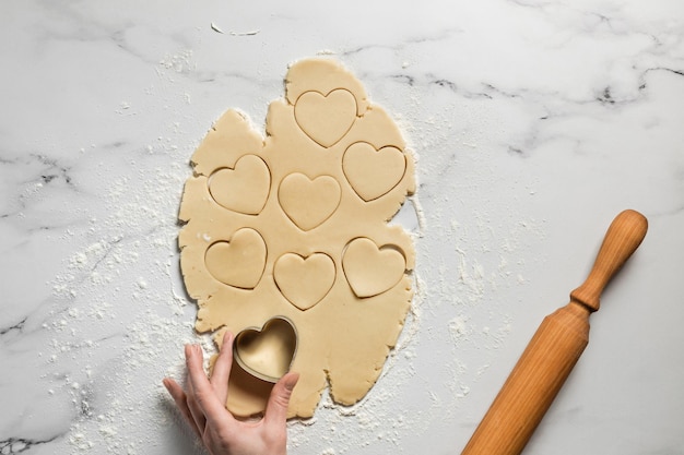 Donna che taglia i biscotti a forma di cuore dalla pasta di pasta frolla stesa sul tavolo di marmo spolverata di mattarello di farina vista dall'alto Cottura per san valentino