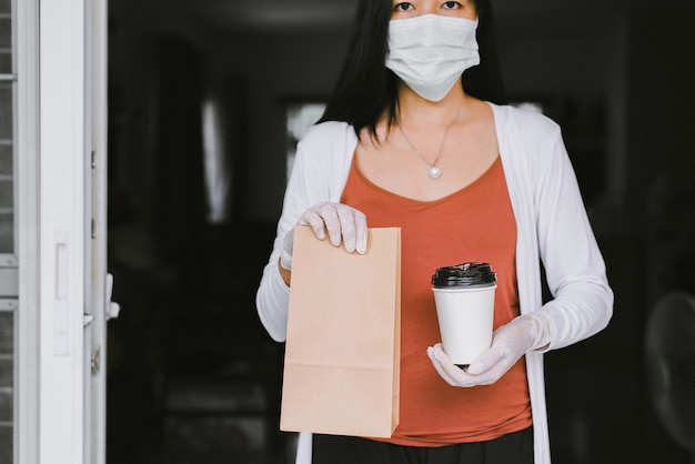 Donna che tiene cibo da asporto in un sacchetto di carta e una tazza di caffè dal servizio di consegna durante la situazione di pandemia di coronavirus a casa