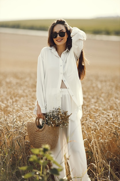 Donna che tiene il mazzo di fiori di campo in un sacchetto di paglia, camminando nel campo di grano. Donna castana che cammina nel campo estivo indossando abiti bianchi