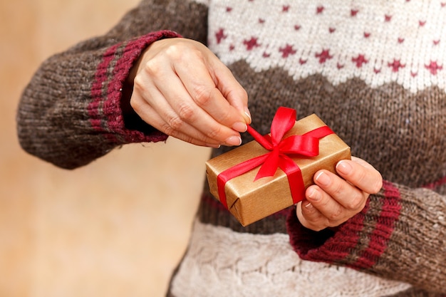 Donna che tiene in mano una confezione regalo con un nastro rosso e cerca di aprirla. Profondità di campo, messa a fuoco selettiva sulla scatola. Concetto di fare un regalo in vacanza o compleanno.