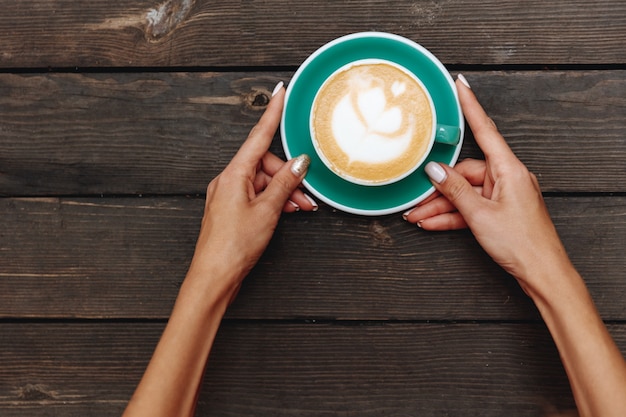 Donna che tiene in mano una tazza verde chiaro di caffè aromatizzato fresco e caldo con latte art a forma di cuore su un tavolo di legno scuro
