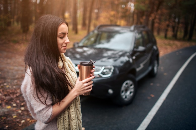 Donna che tiene tazza di caffè eliminabile accanto all'automobile. Primo piano a mano. Concetto di autunno. Viaggio nella foresta d'autunno in auto